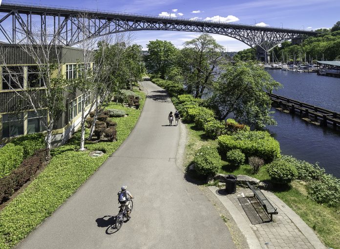 View of Street End from Fremont Bridge A photo of a trail with a cyclist riding on it next to a canal with a bench overlooking it and a bridge in the distance.