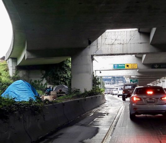 Inslee Pushes Legislation Prioritizing Homeless Encampment Removal Near Highways a photo of a freeway with cars and a tent behind a concrete barrier.