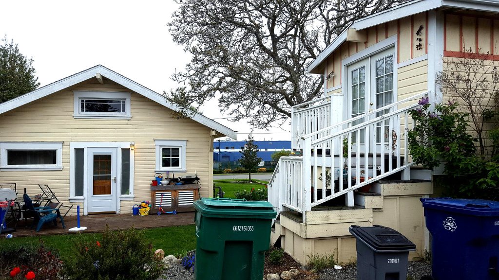 A canary yellow backyard cottage next to house of the same color. Lots of toys and furniture in the lawn shows a family is getting good use out of the space.