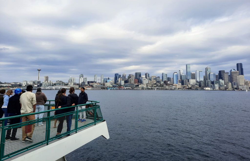 A crowd of people stand on a deck of a ferry as it approaches Seattle