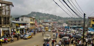 A photo of a dirt street crowded with people and vehicles with a green mountain in the distance and electrical wire