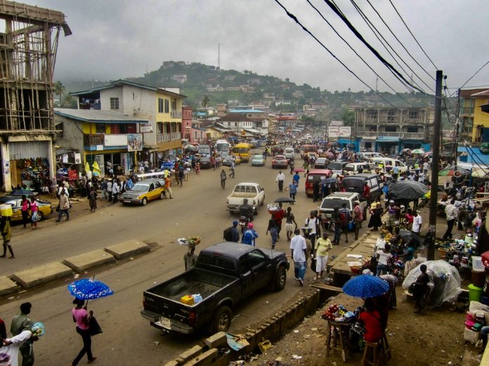 A photo of a dirt street crowded with people and vehicles with a green mountain in the distance and electrical wire