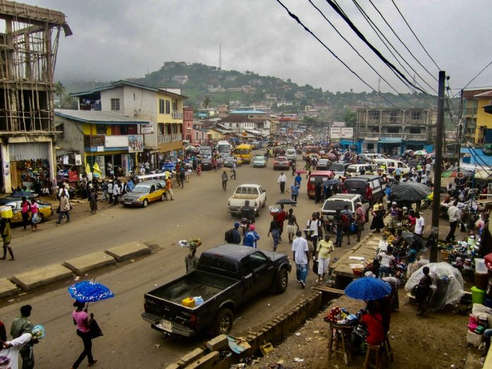 294B6E77-92A8-4248-A868-B365B488EF01_1_201_a A photo of a dirt street crowded with people and vehicles with a green mountain in the distance and electrical wire