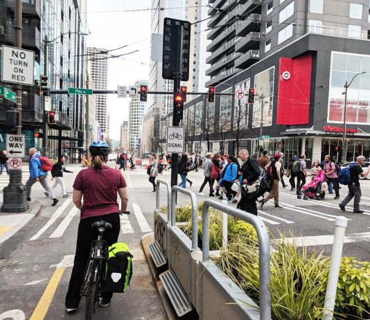 Elected Leaders Shy Away From Big Amendments to 2050 Regional Transportation Plan A bike rider waits at a busy intersection in downtown Seattle as pedestrians cross