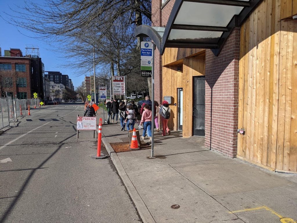 A six foot sidewalk bursting with people walking and some cones and tree pits