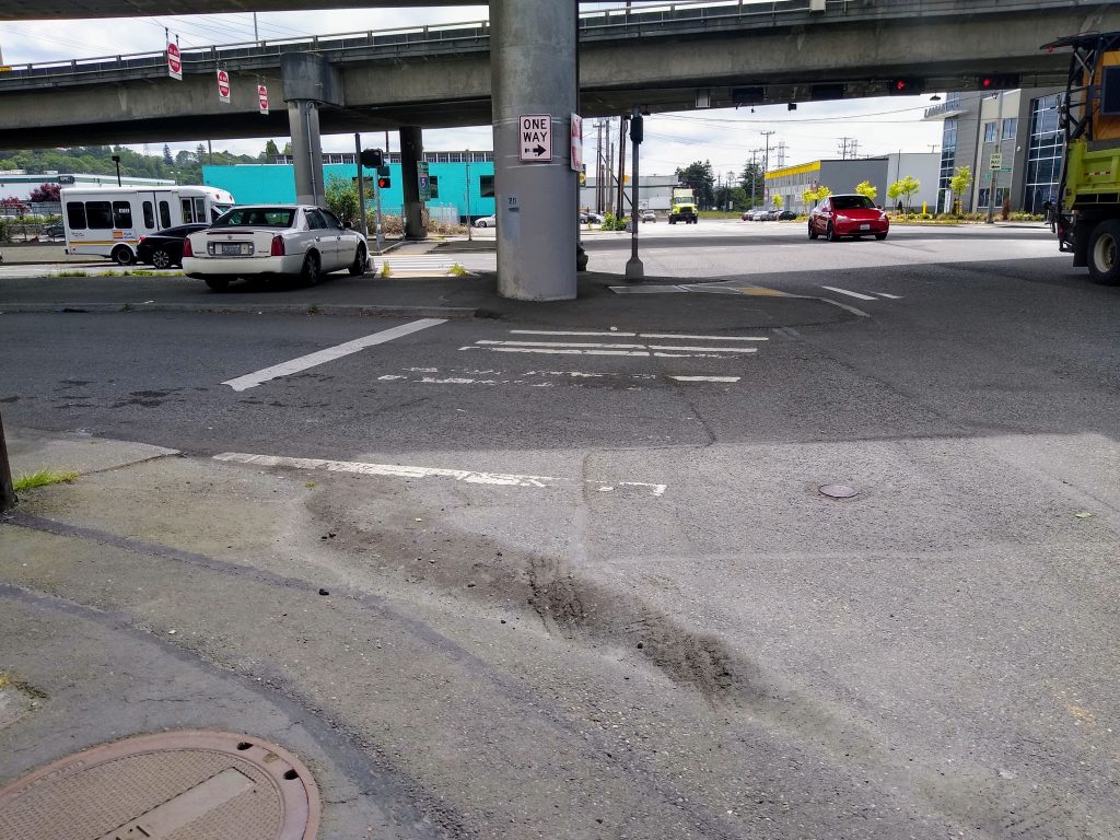 A slanted crosswalk mostly ground off underneath an unpleasant highway overpass with a random car parked on the median