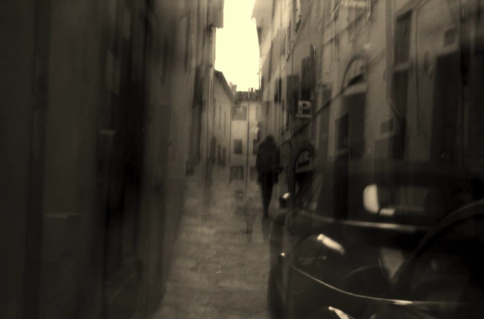 A blurred black and white photo of a figure walking down a street and a car.