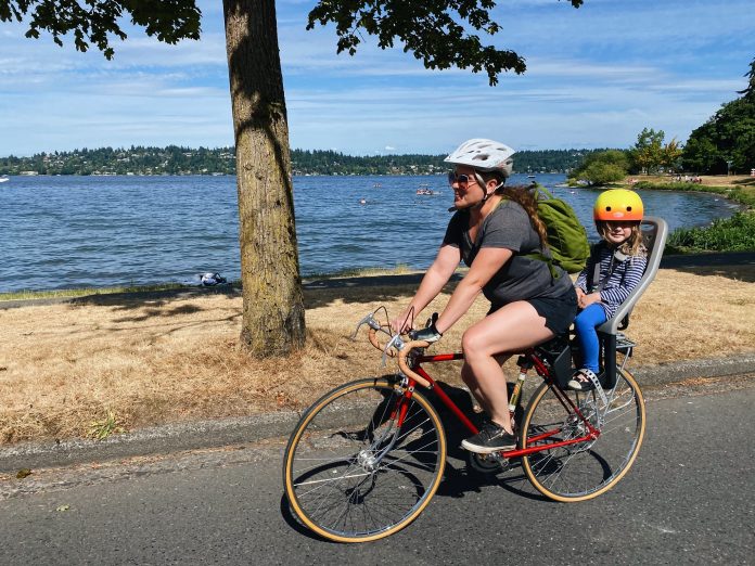 a mother bikes with her chair in the rear on a street near a lake