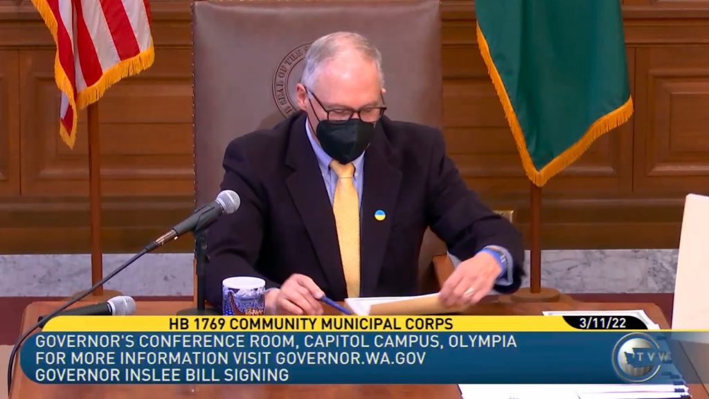 A screen shot from a video showing a man wearing a mask sitting at a desk and reviewing a stack of papers. 