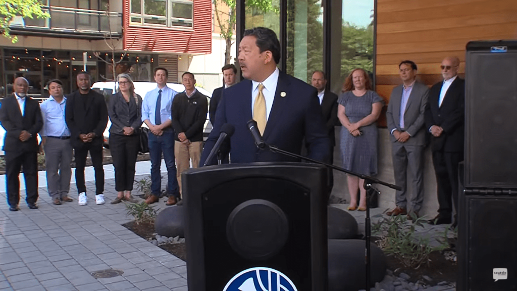Bruce Harrell wears a suit and stands at a podium with a group of people standing behind him.