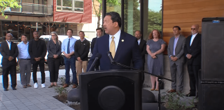 Bruce Harrell wears a suit and stands at a podium with a group of people standing behind him.