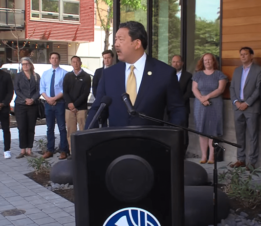 Seattle Leaders Eye JumpStart Housing Funds to Close $230 Million Budget Hole Bruce Harrell wears a suit and stands at a podium with a group of people standing behind him.