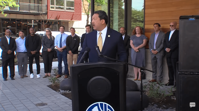 Screenshot (3)-min Bruce Harrell wears a suit and stands at a podium with a group of people standing behind him.