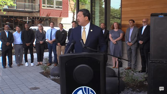 Bruce Harrell wears a suit and stands at a podium with a group of people standing behind him.