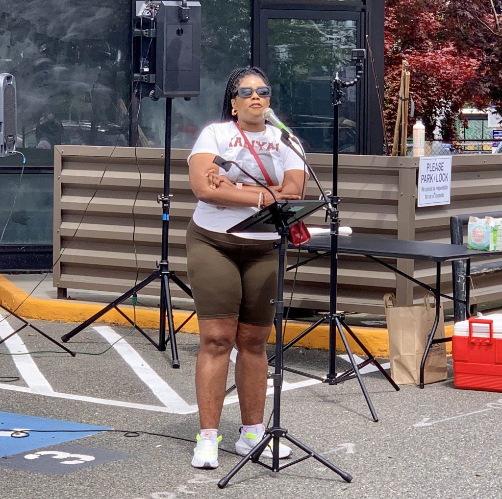 A Black woman wearing sunglasses and a red purse speaks at a podium. 