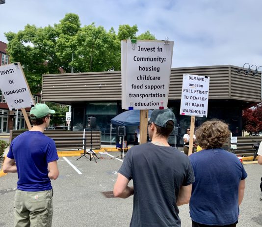 Amazon Walks Away from Warehouse Plans Near Mount Baker Light Rail Station people stand in a parking lot holding stands calling for more community investment