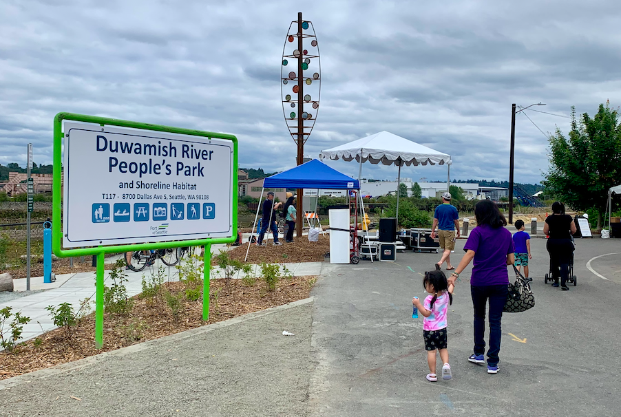 A woman and young girl walk holding hands near the sign for the Duwamish People's Park