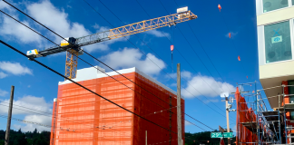 A construction crane over a building under construction with another building under construction across the street and workers standing between them.