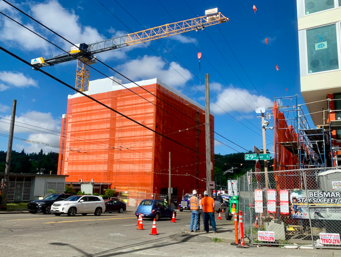 A construction crane over a building under construction with another building under construction across the street and workers standing between them.