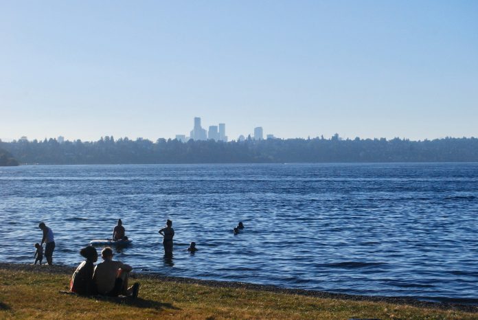 people sit on the grass and splash in the water of Lake Washington with the Seattle skyline in the distance.