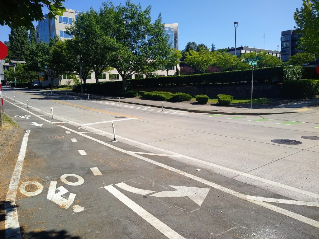 A photo of a bike lane turn with another segment of the facility across the street, with a street sign for SW Andover and 26th Ave SW visible