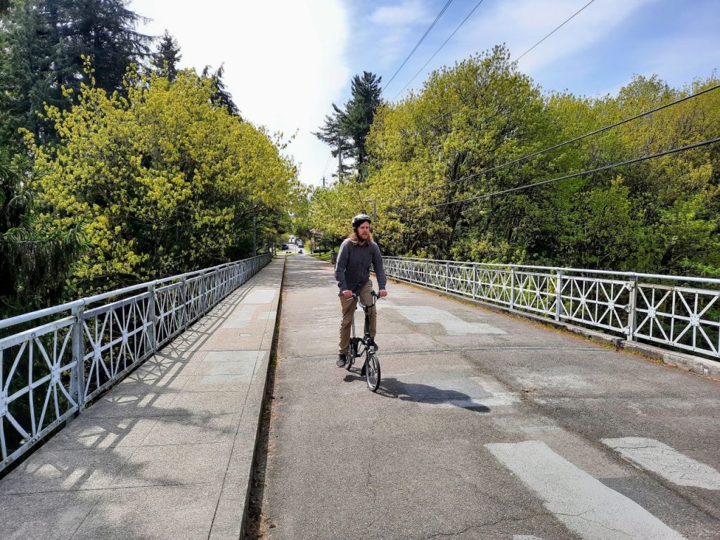 Urbanist writer Ryan Packer rides their bike over the 20th Avenue pedestrianized bridge, with tree canopy on either side.