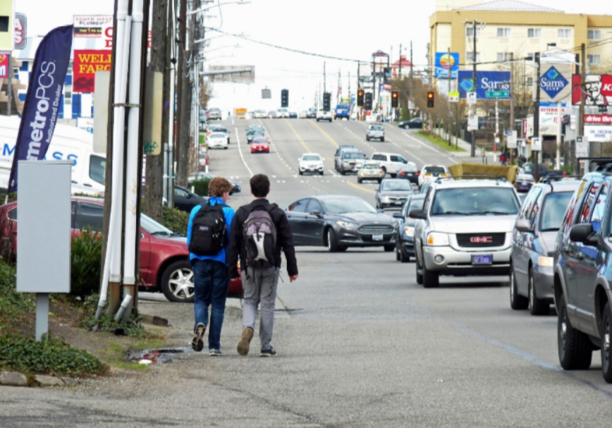 Two school kids walk along a busy state highway with no sidewalks
