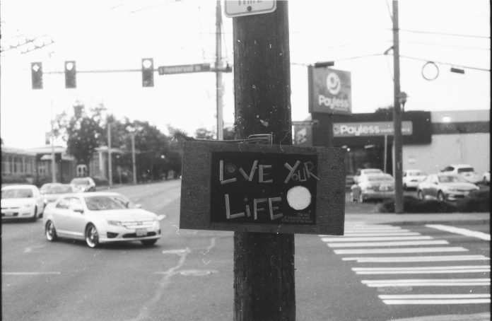 Black and white photo of a large surface intersection and turning car with a telephone pole right in the middle bearing a sign saying "Live Your Life"