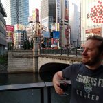 Owen Pickford holding a beer, wearing a Sounders shirt in front of a bridge, river and large towers in Tokyo.