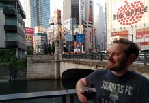Seattle Needs a Powerful Urbanist Media Outlet Owen Pickford holding a beer, wearing a Sounders shirt in front of a bridge, river and large towers in Tokyo.