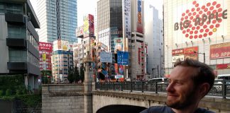 Owen Pickford holding a beer, wearing a Sounders shirt in front of a bridge, river and large towers in Tokyo.