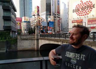Seattle Needs a Powerful Urbanist Media Outlet Owen Pickford holding a beer, wearing a Sounders shirt in front of a bridge, river and large towers in Tokyo.