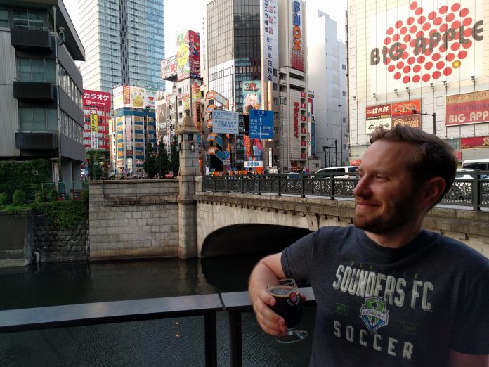 Owen Pickford holding a beer, wearing a Sounders shirt in front of a bridge, river and large towers in Tokyo.