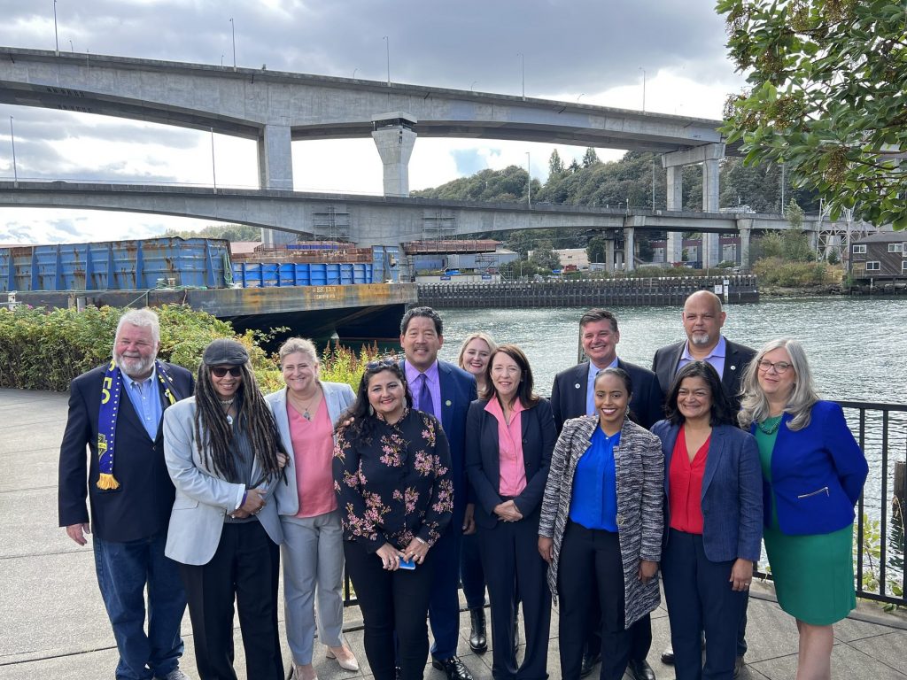 A bunch of elected officials pose for a photo in front of the West Seattle bridge from harbor island