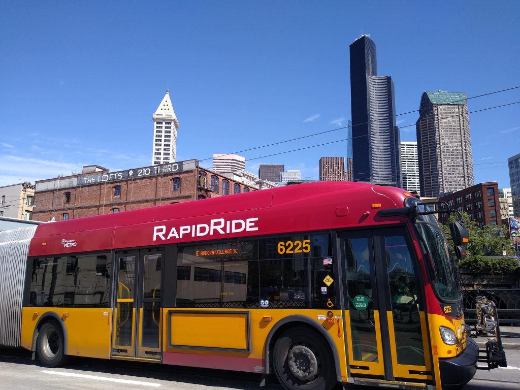 A red and gold RapidRide bus in Pioneer Square with the Seattle skyline in the background.