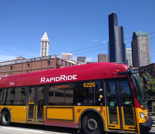 County Council Grapples With Future of King County Metro in Proposed Budget A red and gold RapidRide bus in Pioneer Square with the Seattle skyline in the background.