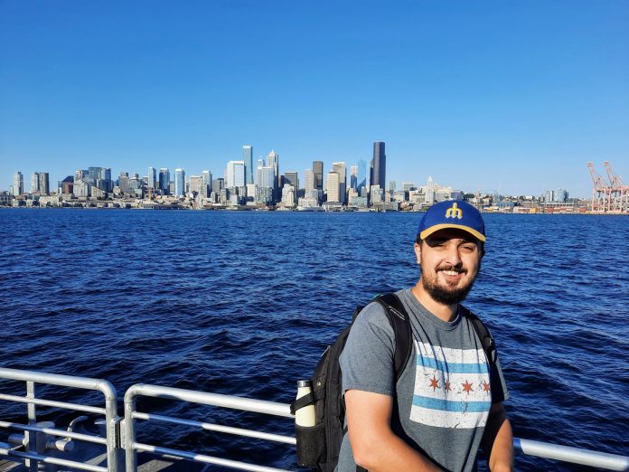 Rian Watt, pictured here with Elliott Bay and the Seattle Skyline. He wears a Mariners hat.