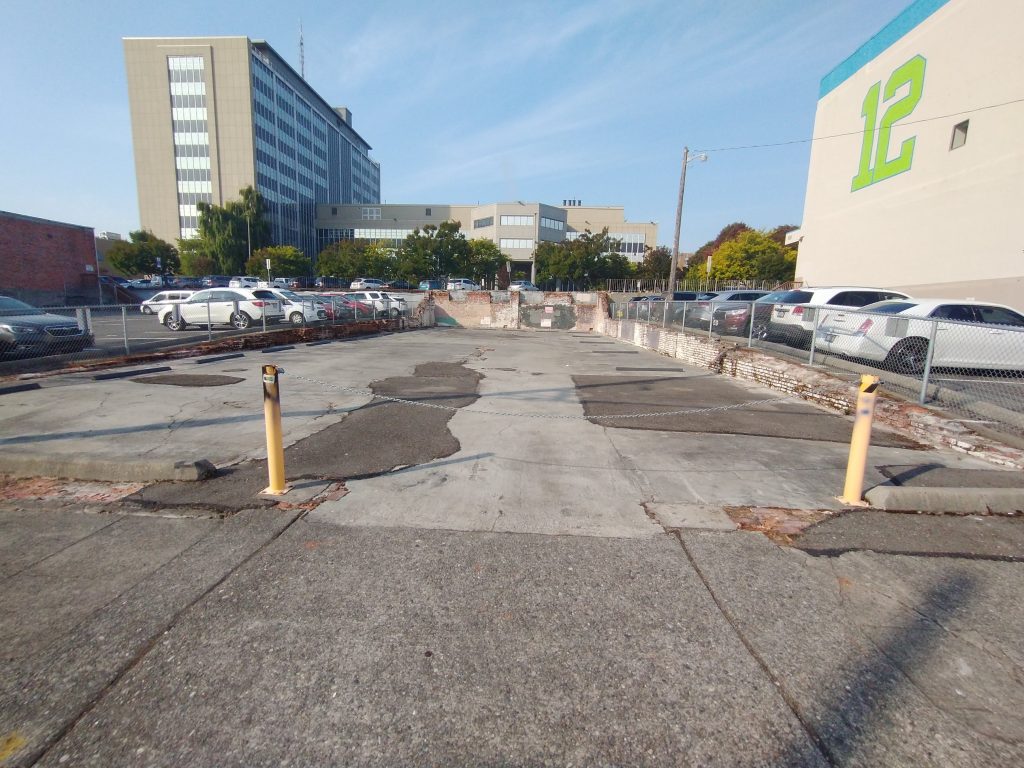A photo of an empty lots surrounding by surface level parking and large buildings.