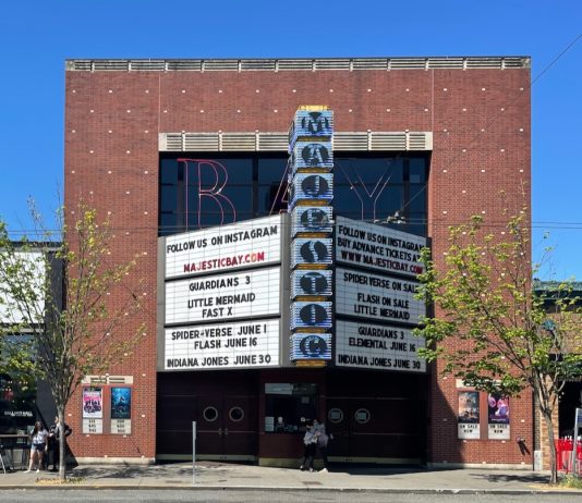 Your 2023 Urbanist Summer Movie List Street scape brick facade movie theater building in front of a blue sky featuring a marquee advertising Majestic Bay, Guardians 3, Little Mermaid, Fast X and coming attractions of Indiana Jones and Spiderverse.
