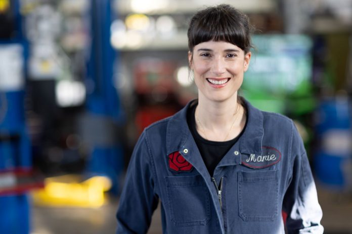 A young dark-haired woman in her blue autobody shop overalls.