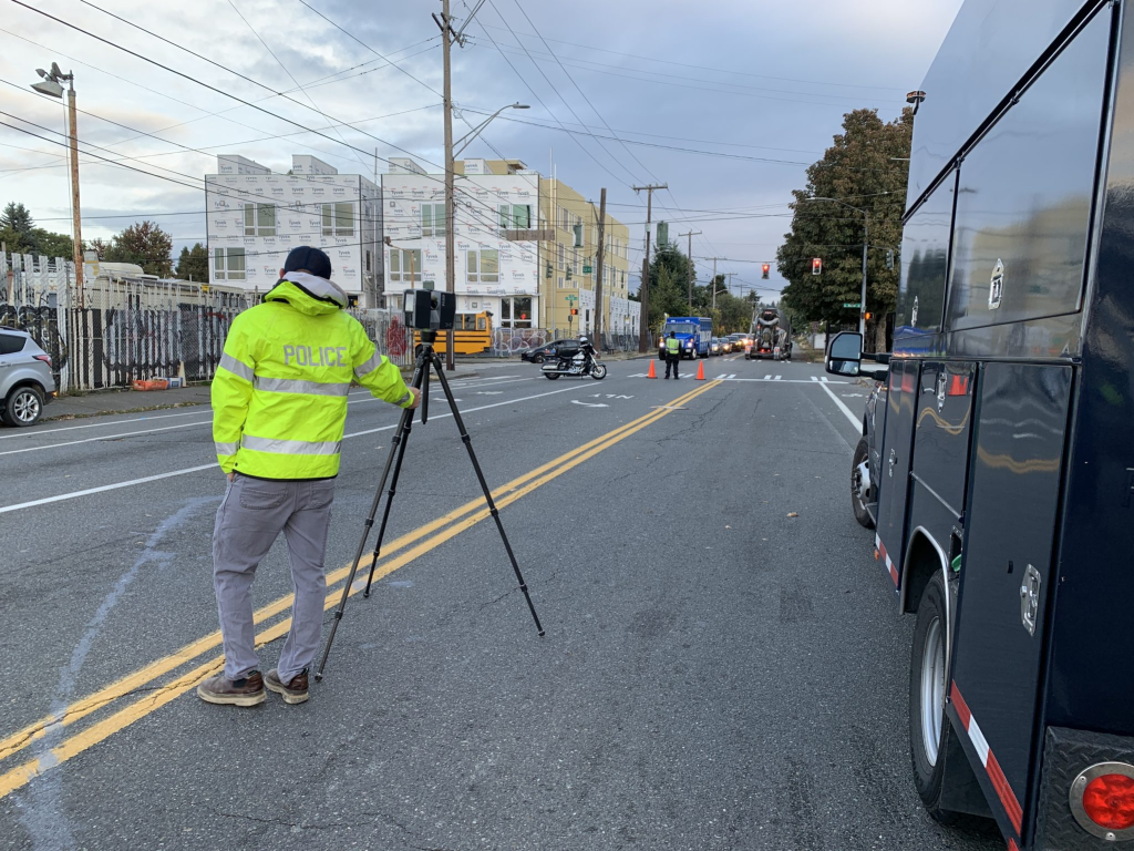 A police officer stands in the road with a camera in advance of an intersection where a crash has occurred