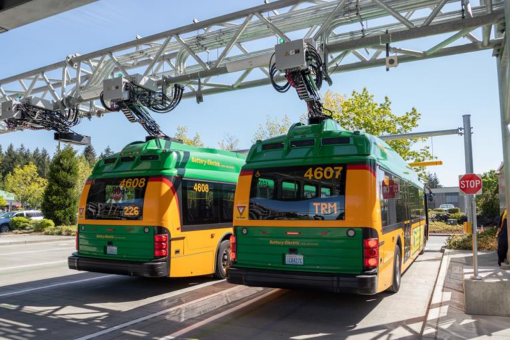 Two bright orange and green buses get charged at a bus base on a sunny day