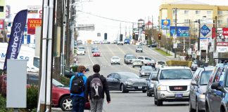 Rows of cars on a block of drive thrus and strip malls as two kids walk in the gutter of the road.