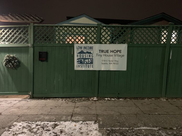 A sign for True Hope Tiny House Village on a green fence in front of a snowy sidewalk.