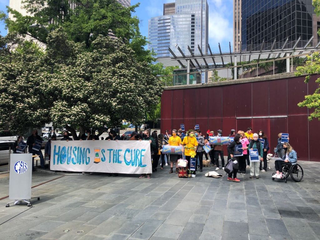 A Stay Housed rally with a "Housing is the Cure" banner at Seattle City Hall with a crowd of about 50.