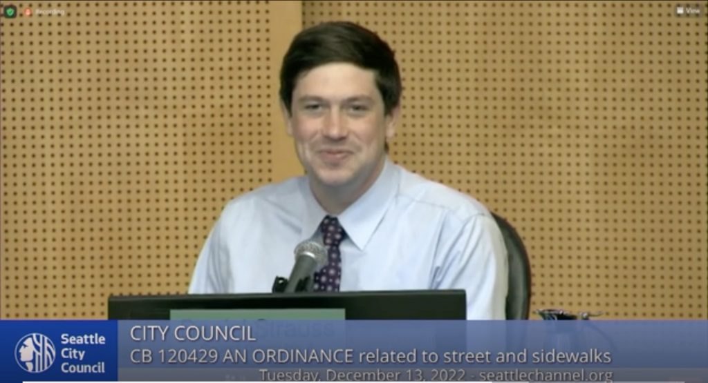 Image description: Strauss wears a boyish smile and a blue tie while sitting at the dais in Council Chambers.