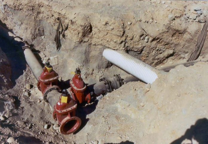 Trench cut in dirt with an intersection of iron pipes and a lightly colored stormwater crossing.