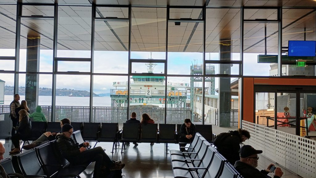 Passengers sit in a new ferry terminal, with a glass wall showing views of a ferry at a dock and Elliott Bay and West Seattle beyond