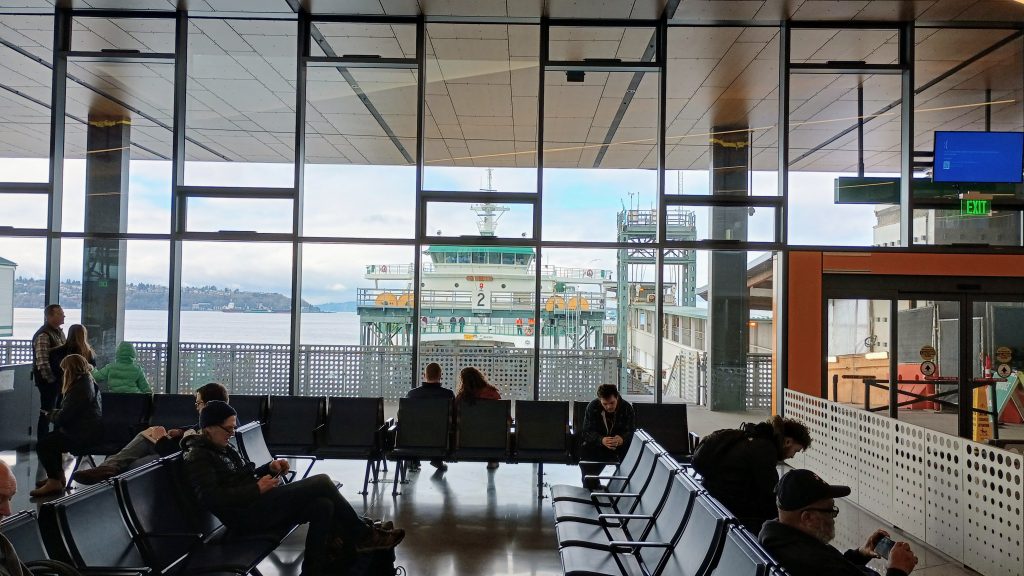 Passengers sit in a new ferry terminal, with a glass wall showing views of a ferry at a dock and Elliott Bay and West Seattle beyond