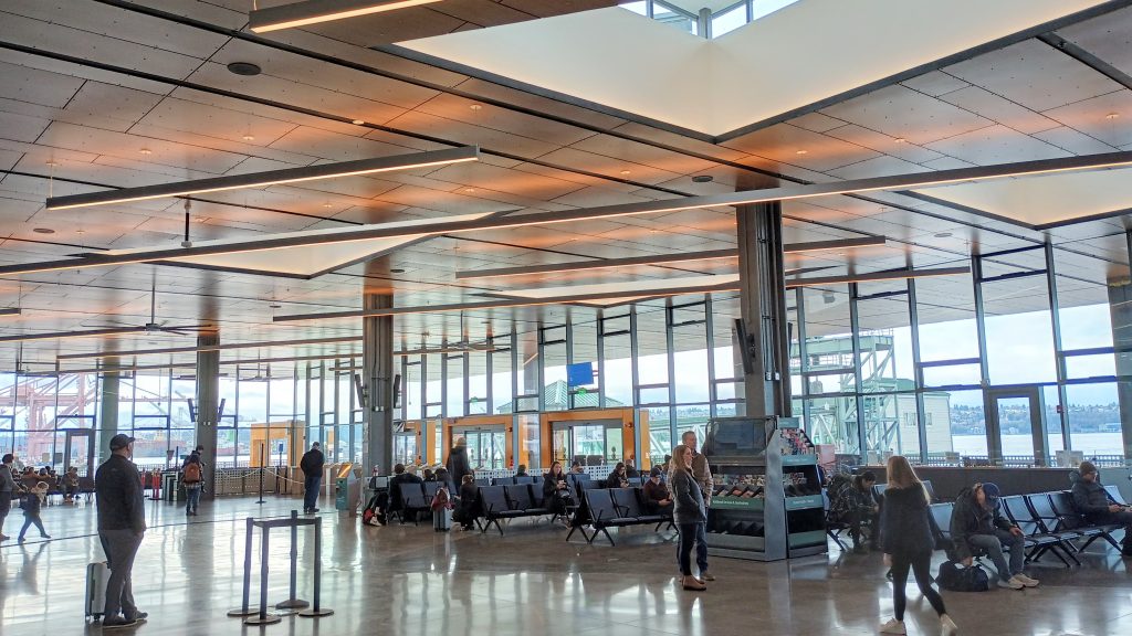 Triangular skylights on the roof of a large ferry terminal with seats, people milling, and a tourist brochure stand